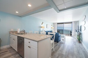 A modern kitchen with a central island, a dining area with white chairs, and a balcony with a view of the ocean.