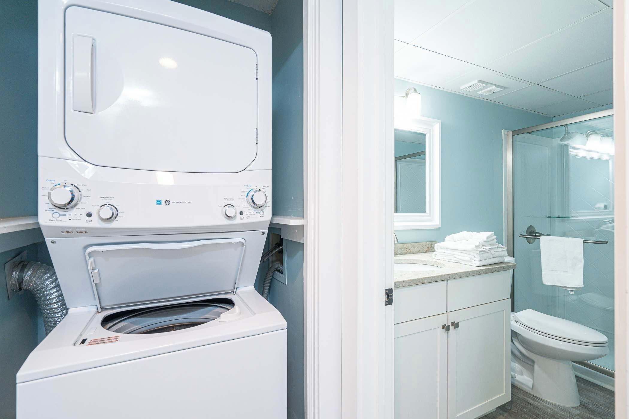 Une petite buanderie équipée d’un lave-linge et d’un sèche-linge à chargement frontal, d’un petit plan de travail avec un miroir et d’une douche à parois de verre.