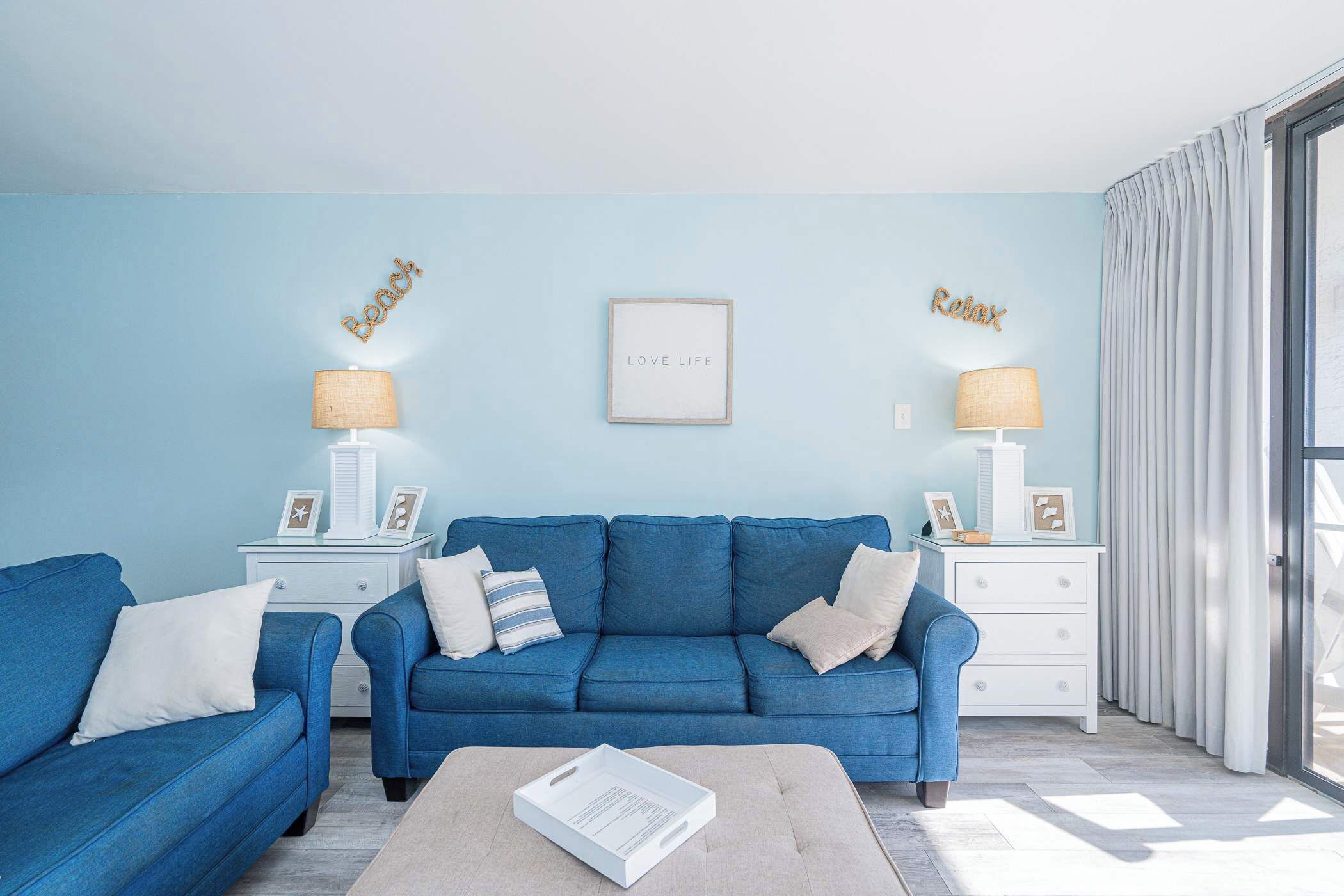A living room with a blue sofa, two side tables with lamps, a framed inspirational quote, and a view through a glass door.