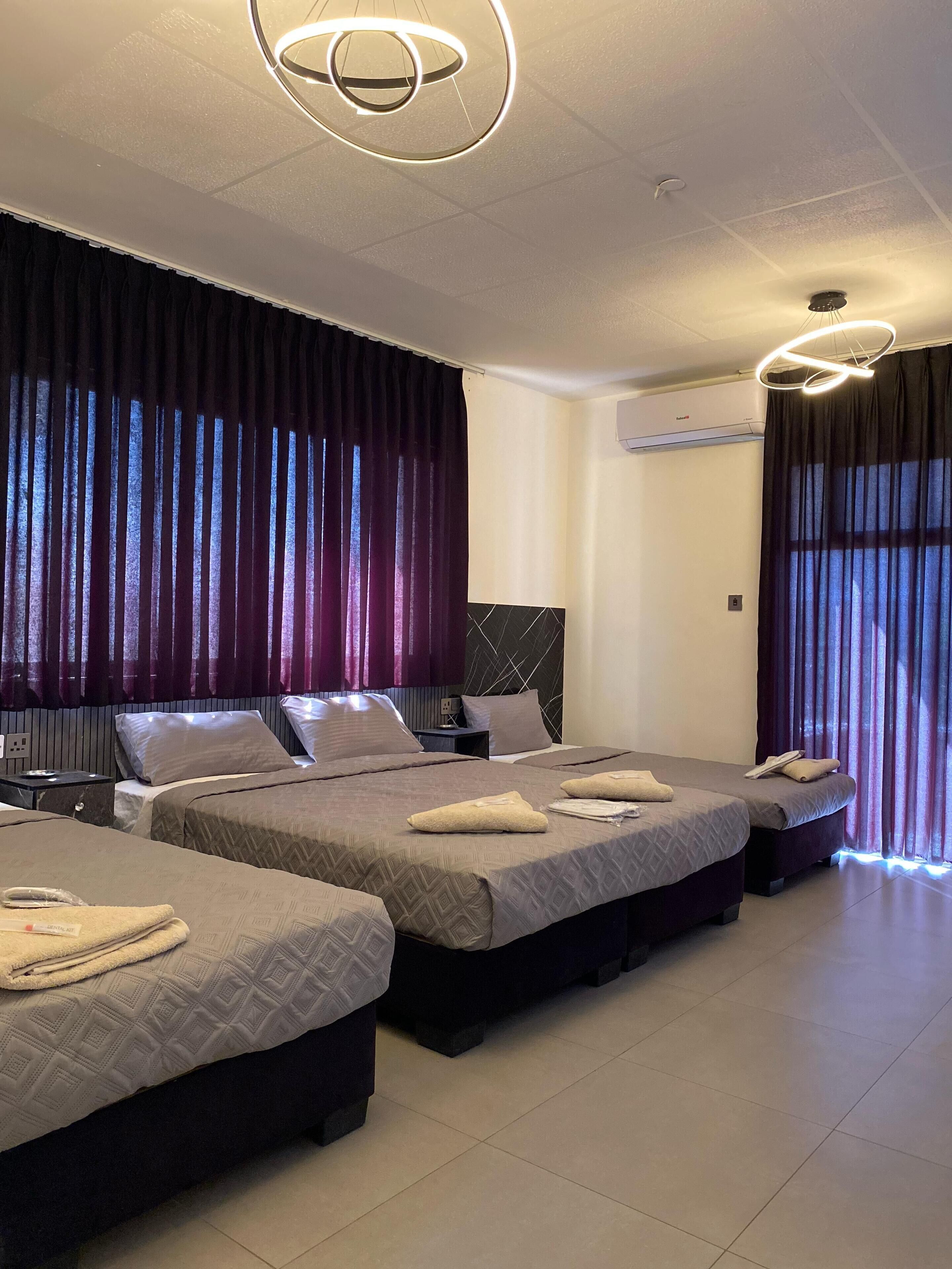 A hotel room with three beds, dark curtains, a modern light fixture, and an air conditioning unit on the wall.