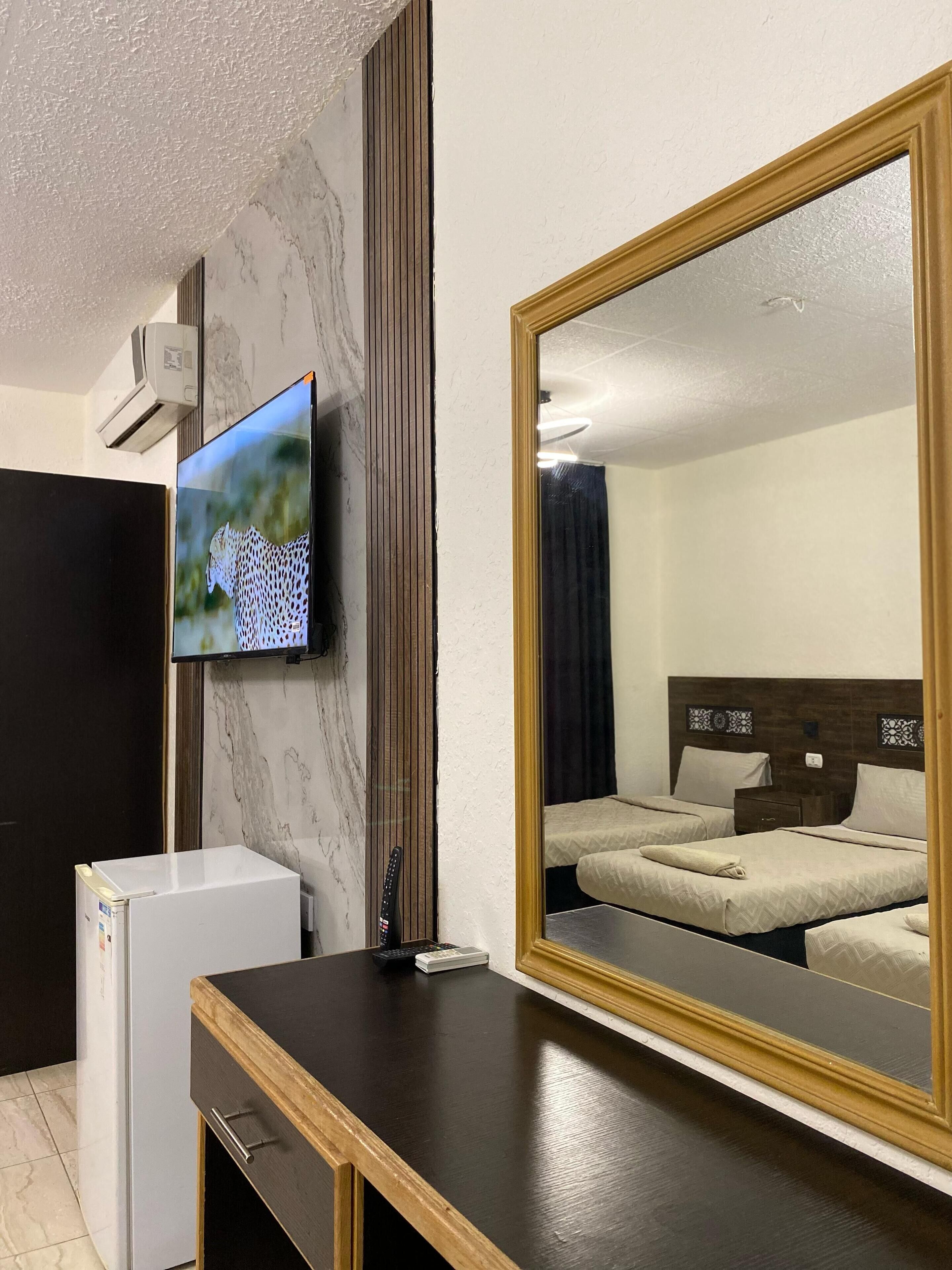 Een hotelkamer met een grote spiegel, een flatscreen televisie aan de wand, een kleine koelkast en een houten bureau.
