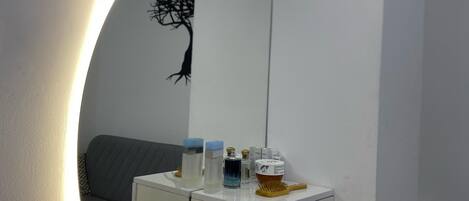 A bathroom with a round mirror, a vanity with toiletries, and a grey sofa in the background.