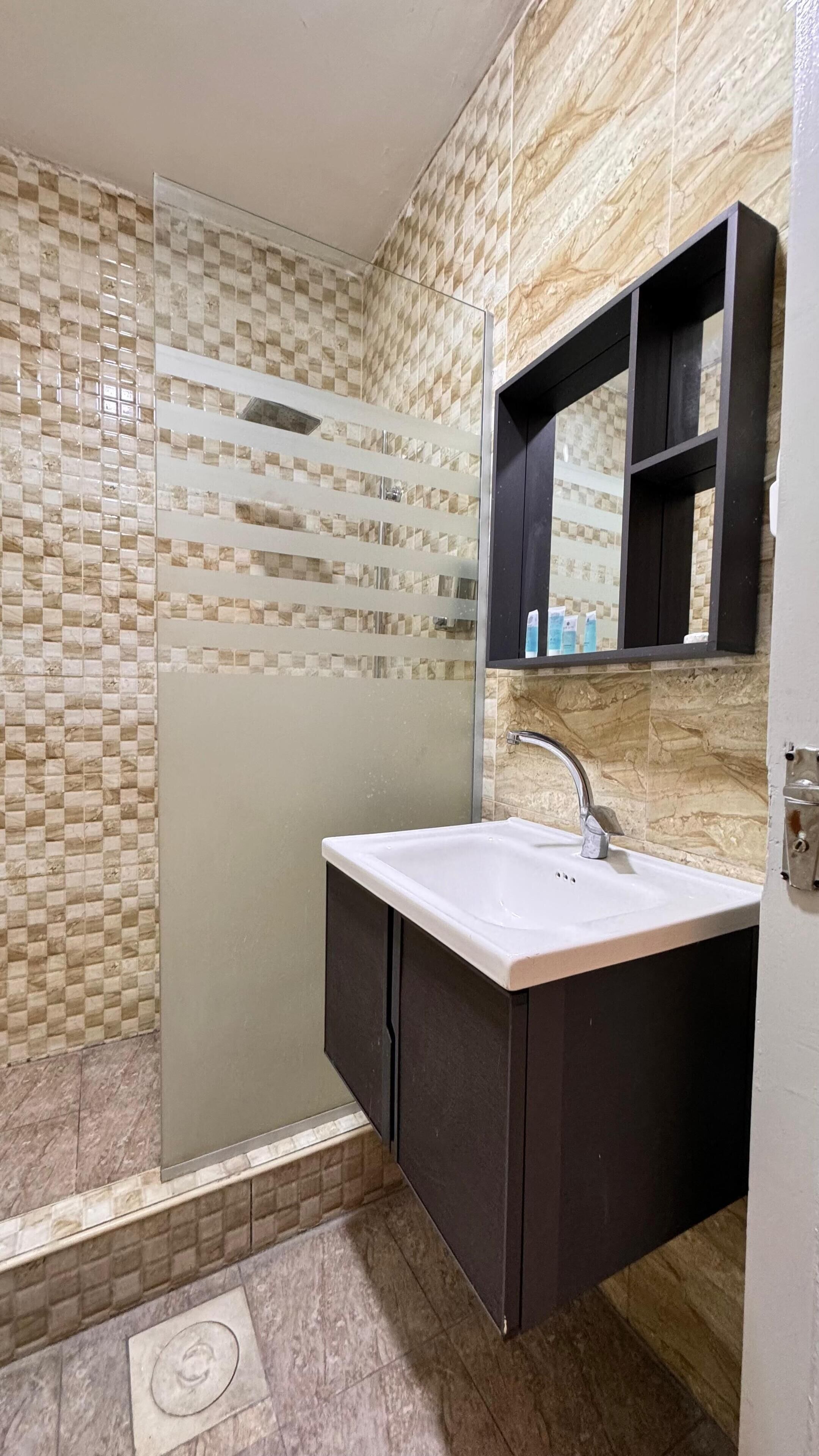 A bathroom with a glass shower enclosure, a white sink, dark cabinets, and a mirror above the sink.