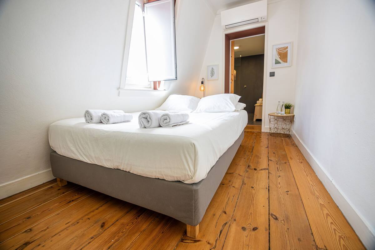 A neatly made bed with rolled towels, a wooden floor, and a small side table with a plant.