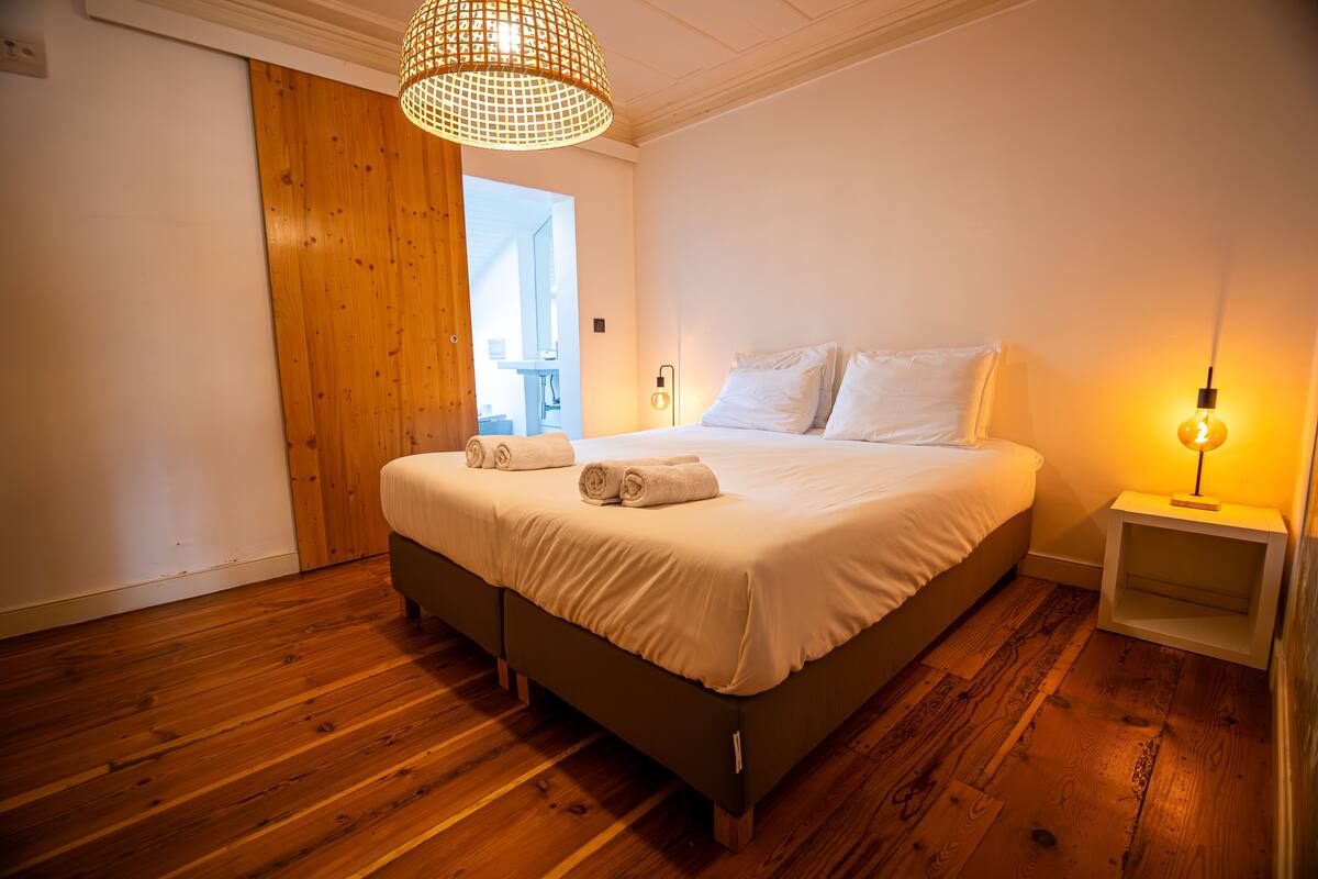 A bedroom with a large bed, wooden flooring, a hanging light fixture, and a nightstand with a lamp.