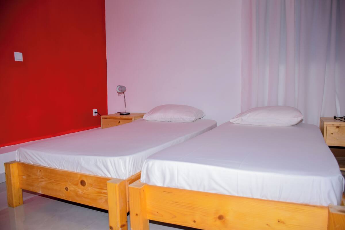 Una habitación con dos camas de madera, ropa de cama blanca, una mesita de noche con lámpara y pared roja.