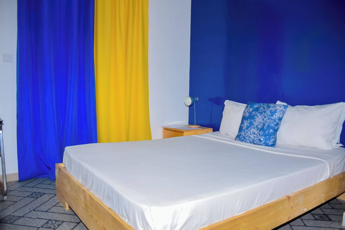 Un dormitorio con cama de madera, ropa de cama blanca, una mesita de noche con lámpara y una cortina azul y amarilla.