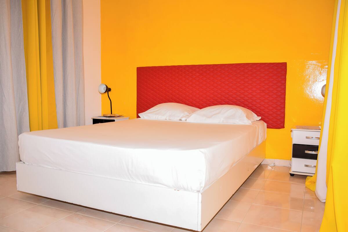 Una cama con ropa de cama blanca, un cabecero rojo y una mesita de noche con una lámpara.