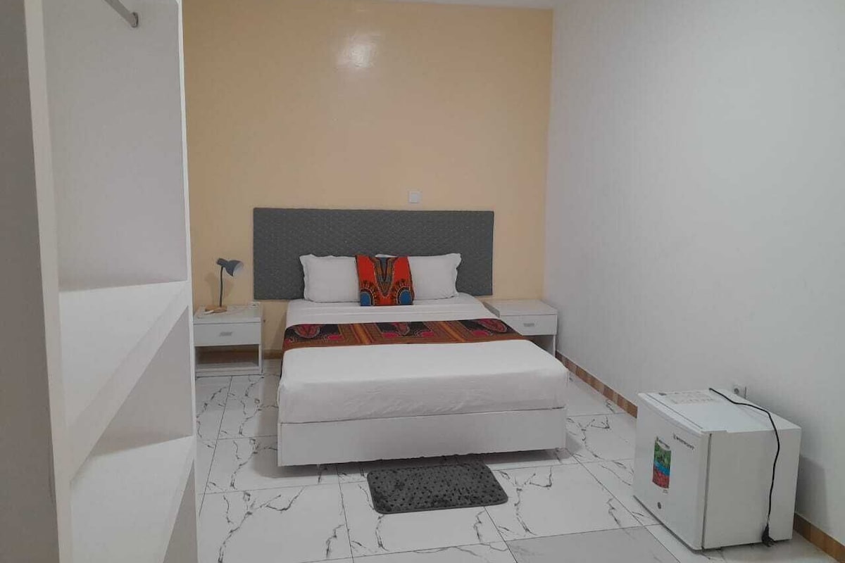 A bedroom with a bed, bedside tables, a refrigerator, and a tiled floor.