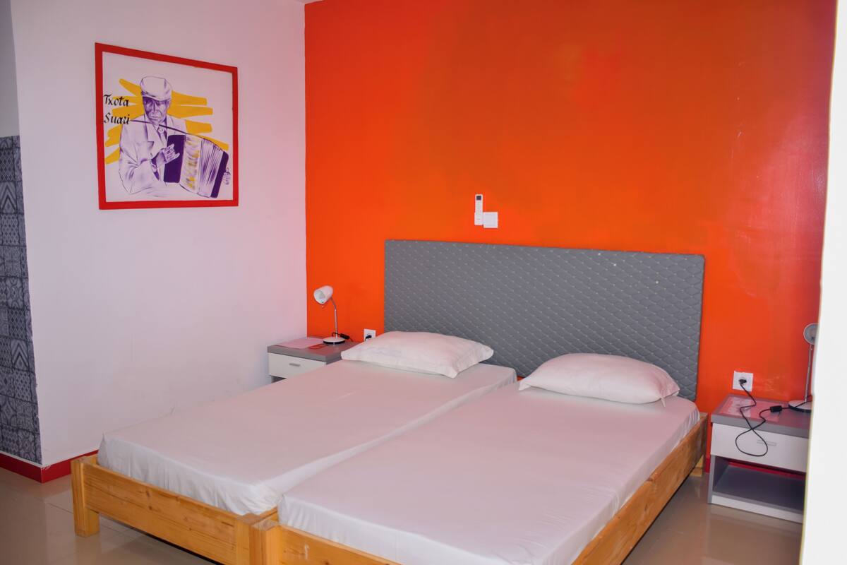 Una cama doble con cabecera gris, dos mesitas de noche y una lámpara fijada a la pared.