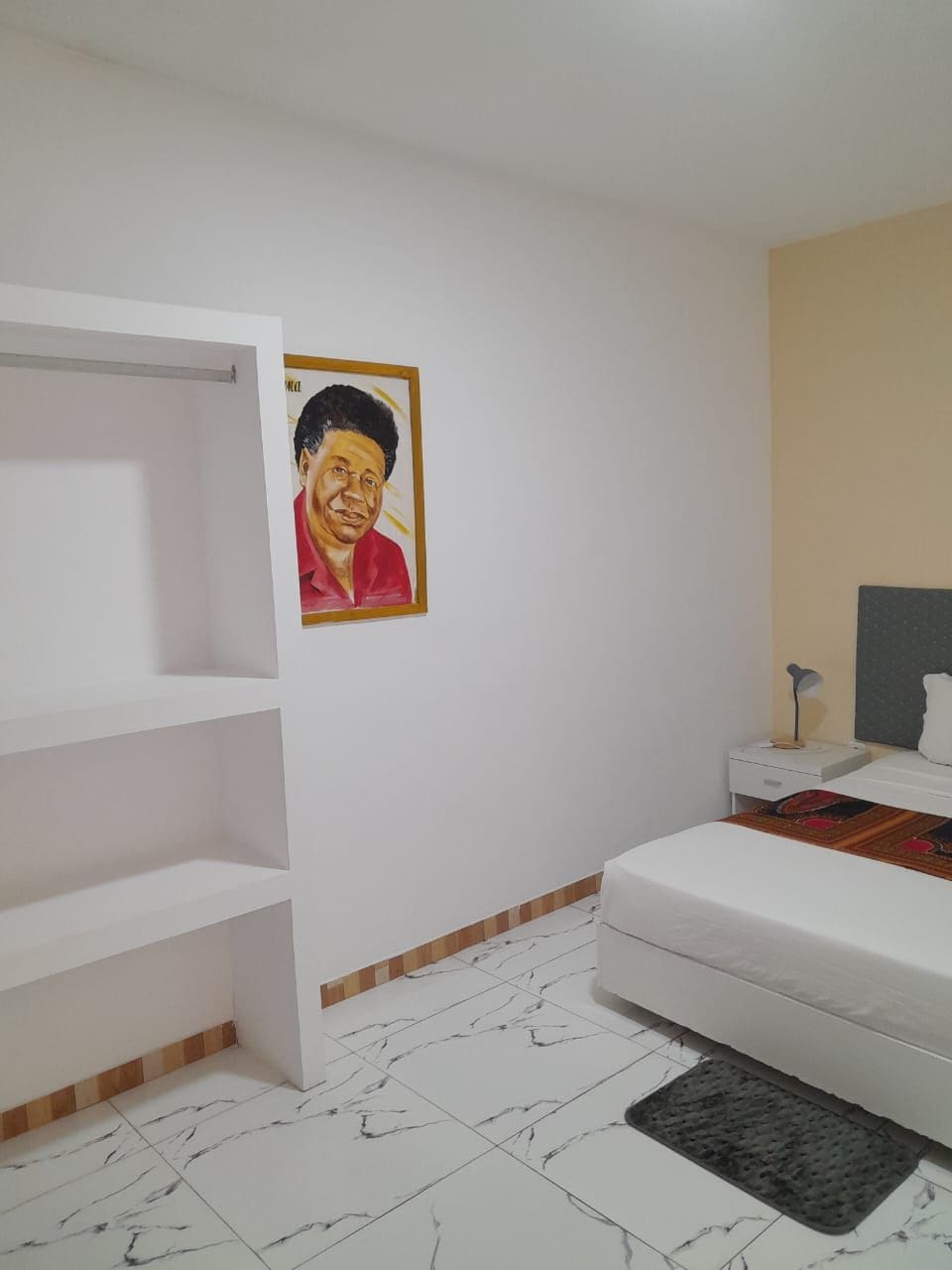 Un dormitorio con cama, mesita de noche, lámpara, estante y un cuadro en la pared.