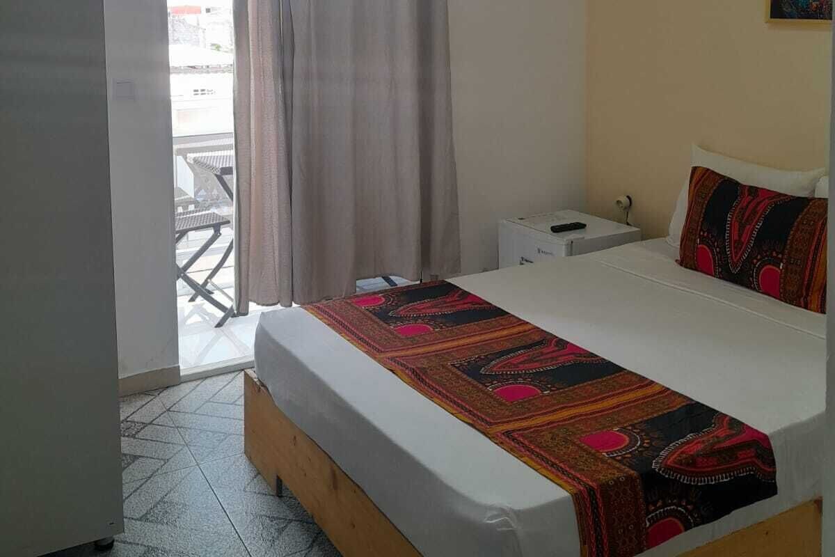 A hotel room with a bed, a bedside table, a nightstand, a painting on the wall, and a view through a window with curtains.