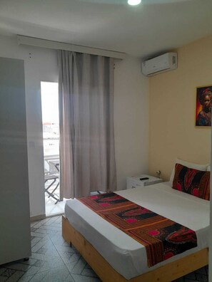 A hotel room with a bed, a bedside table, a nightstand, a painting on the wall, and a view through a window with curtains. - Hotel Principal (Tarrafal)