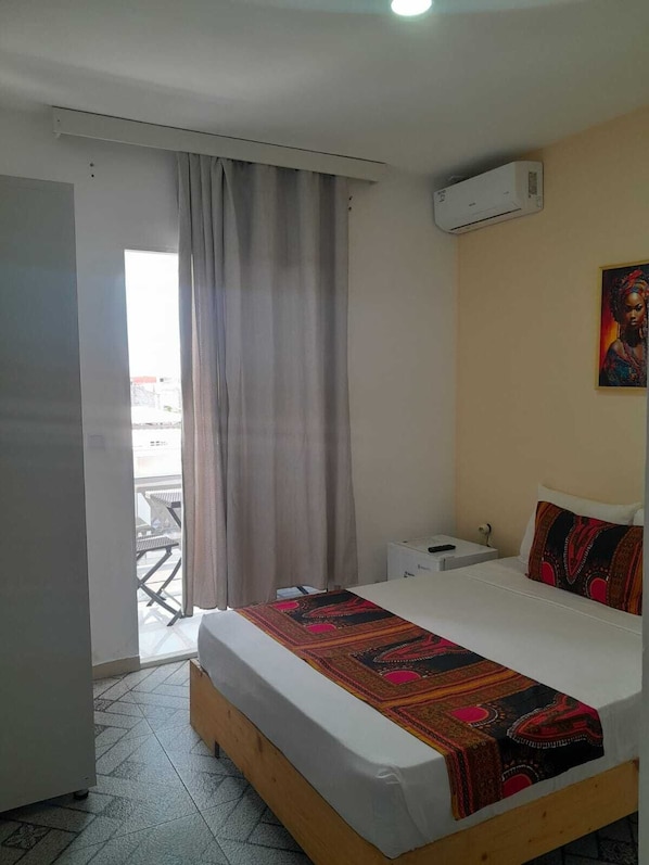 A hotel room with a bed, a bedside table, a nightstand, a painting on the wall, and a view through a window with curtains. - Hotel Principal (Tarrafal)