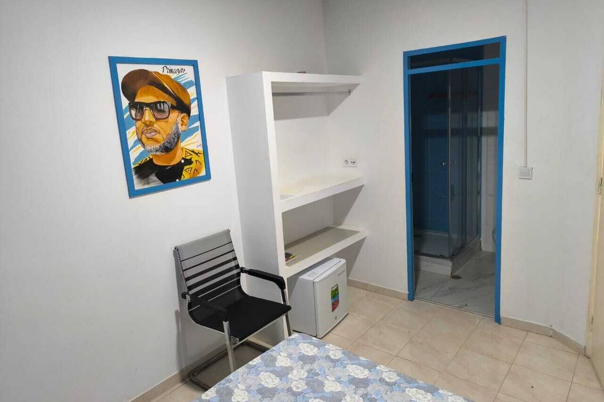 Una habitación con literas, un escritorio con silla, una mesita con mantel floreado, un cartel en la pared y una puerta con marco azul.
