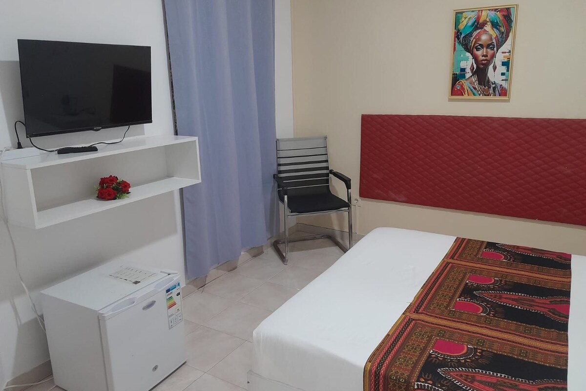 A bedroom with a bed, a television on a stand, a chair, a small refrigerator, and a wall-mounted red headboard.