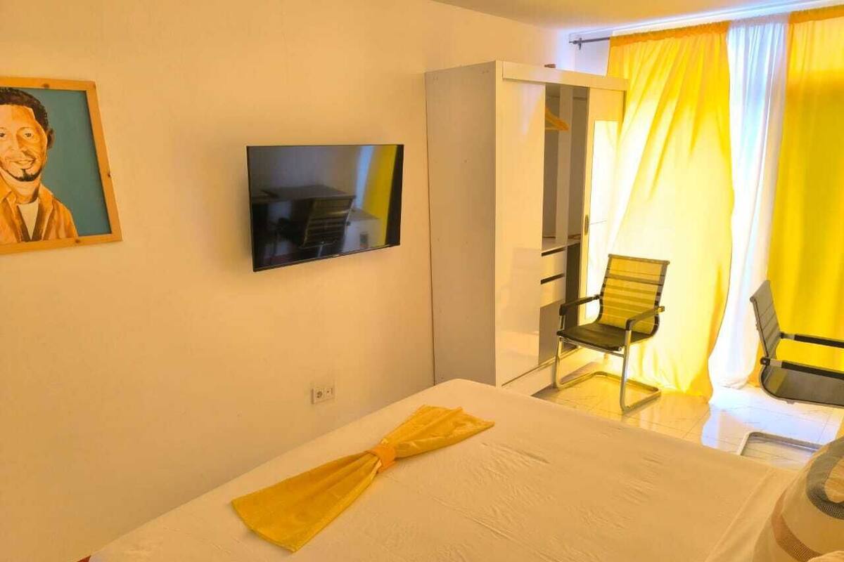 Habitación de hotel con cama, televisor montado en la pared, una silla y una ventana con cortinas amarillas.