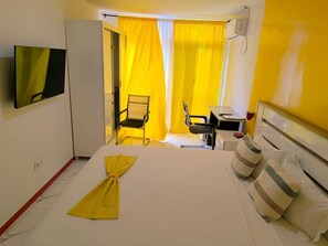 A hotel room with a bed, a desk, a chair, a television, and a window with yellow curtains. - Hotel Principal (Tarrafal)