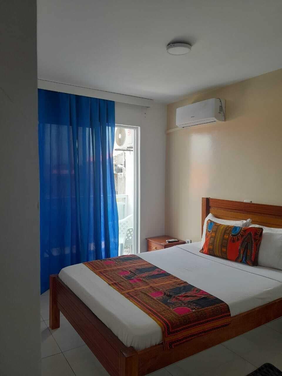 Une chambre à coucher avec un lit, une table de chevet, une fenêtre avec des rideaux bleus et un appareil de climatisation fixé au mur.