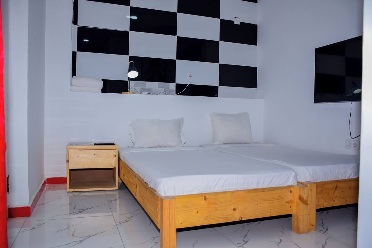 Un dormitorio con cama de madera, ropa de cama blanca, una mesita de noche pequeña, un televisor montado en la pared y un diseño de pared a cuadros.