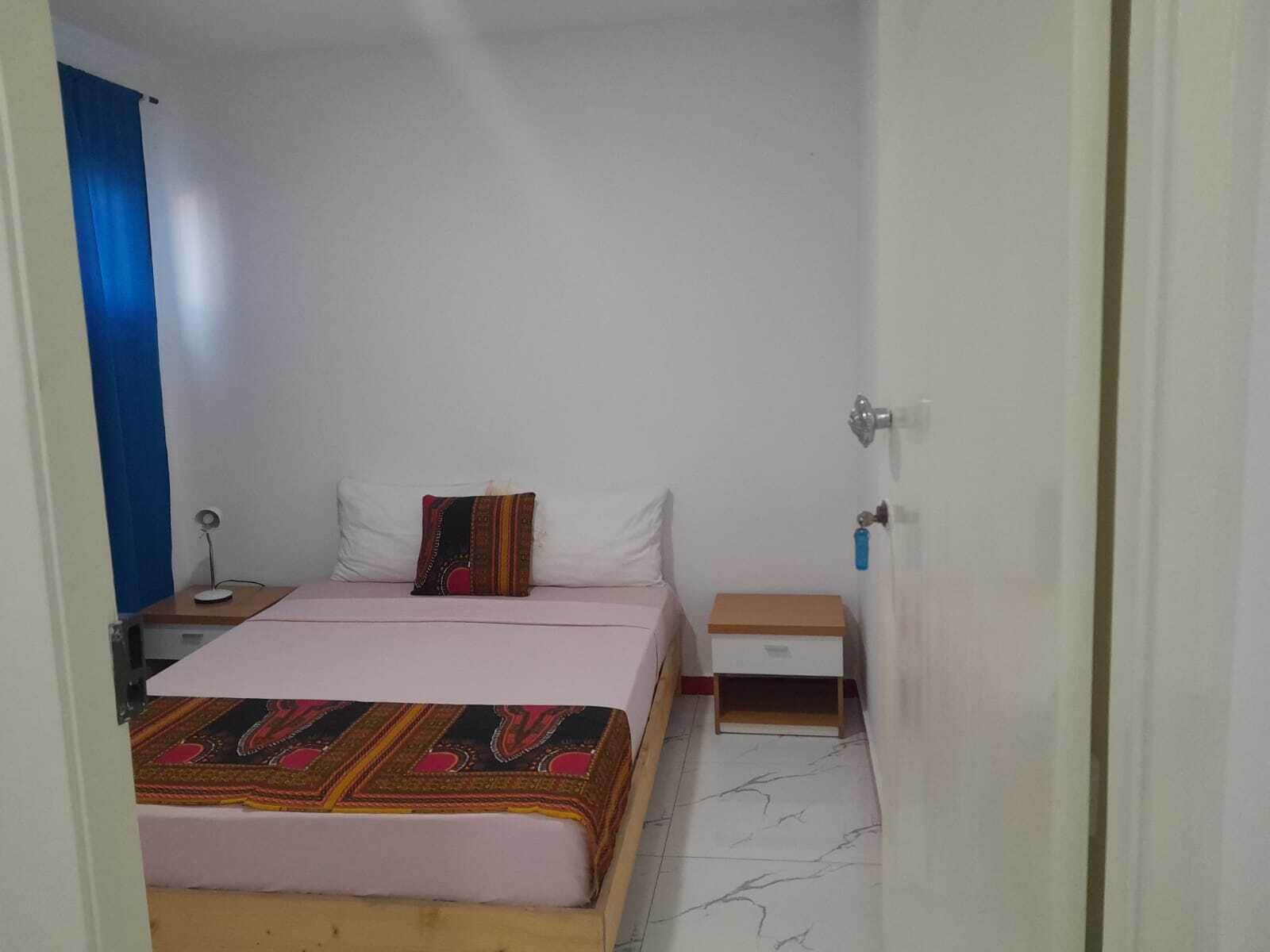 A small, single-bed room with a wooden headboard, a bedside table with a lamp, a small drawer unit, and a door with a key lock.