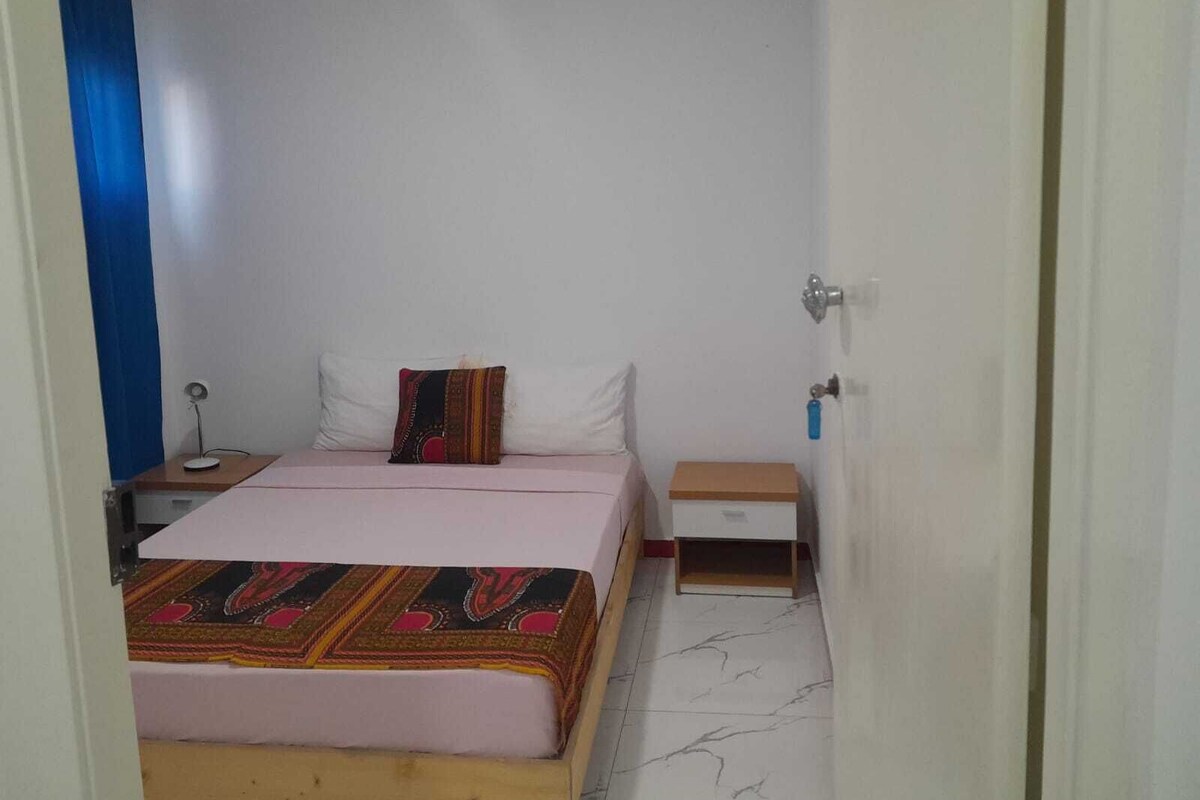 A small, single-bed room with a wooden headboard, a bedside table with a lamp, a small drawer unit, and a door with a key lock.