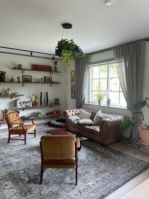 Uma sala de estar com um sofá de couro marrom, uma cadeira de madeira, um tapete estampado e prateleiras com objetos decorativos.