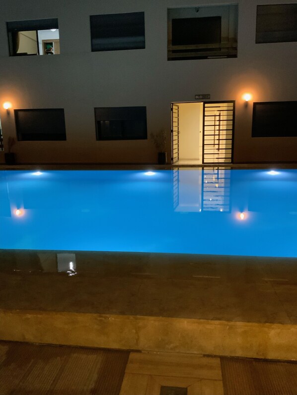 Ein Schwimmbecken mit Fliesenboden und einer umlaufenden Holzterrasse. An den Wänden befinden sich Fenster mit Jalousien, und eine Tür mit einem Metallgitter ist sichtbar.