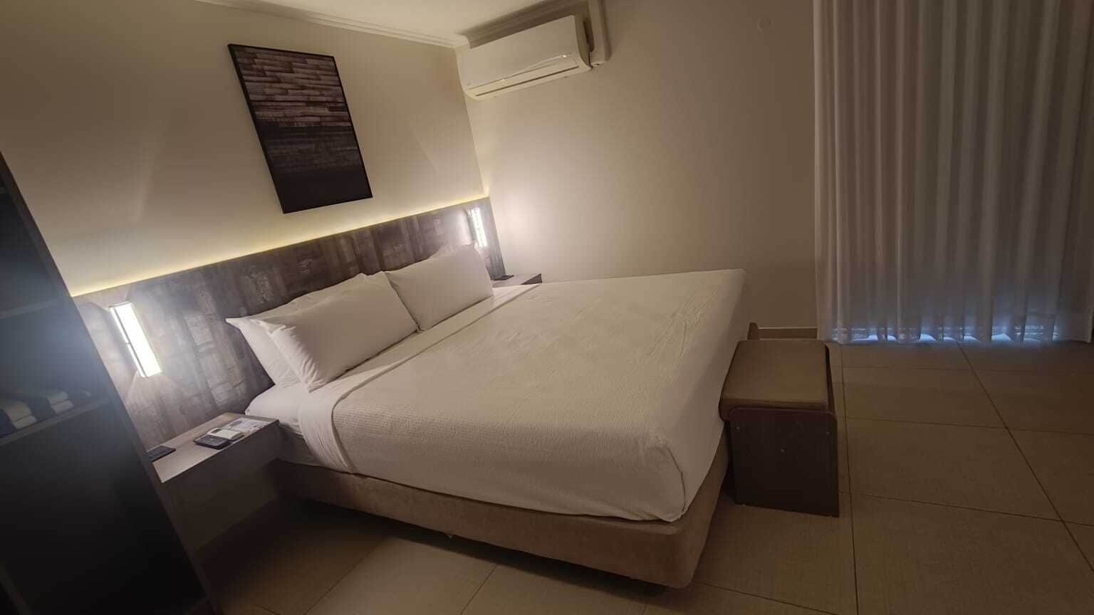 A neatly made bed with white linens, a bedside table with a lamp, and a wall-mounted air conditioning unit.