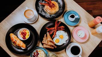 Una mesa de desayuno con diferentes platos, como panqueques, huevos, tocino, tostadas y un croissant.