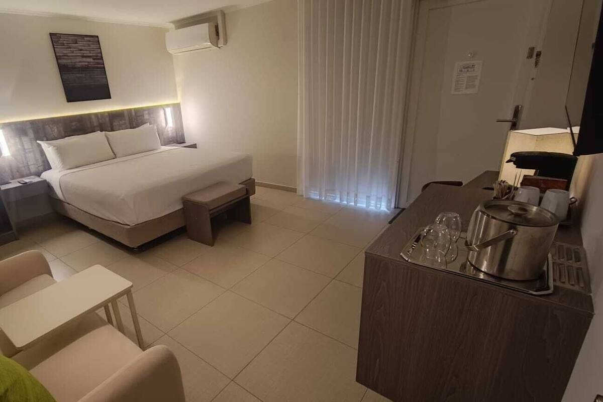 A hotel room with a bed, a desk, a chair, a small table, a minibar, and a window with curtains.