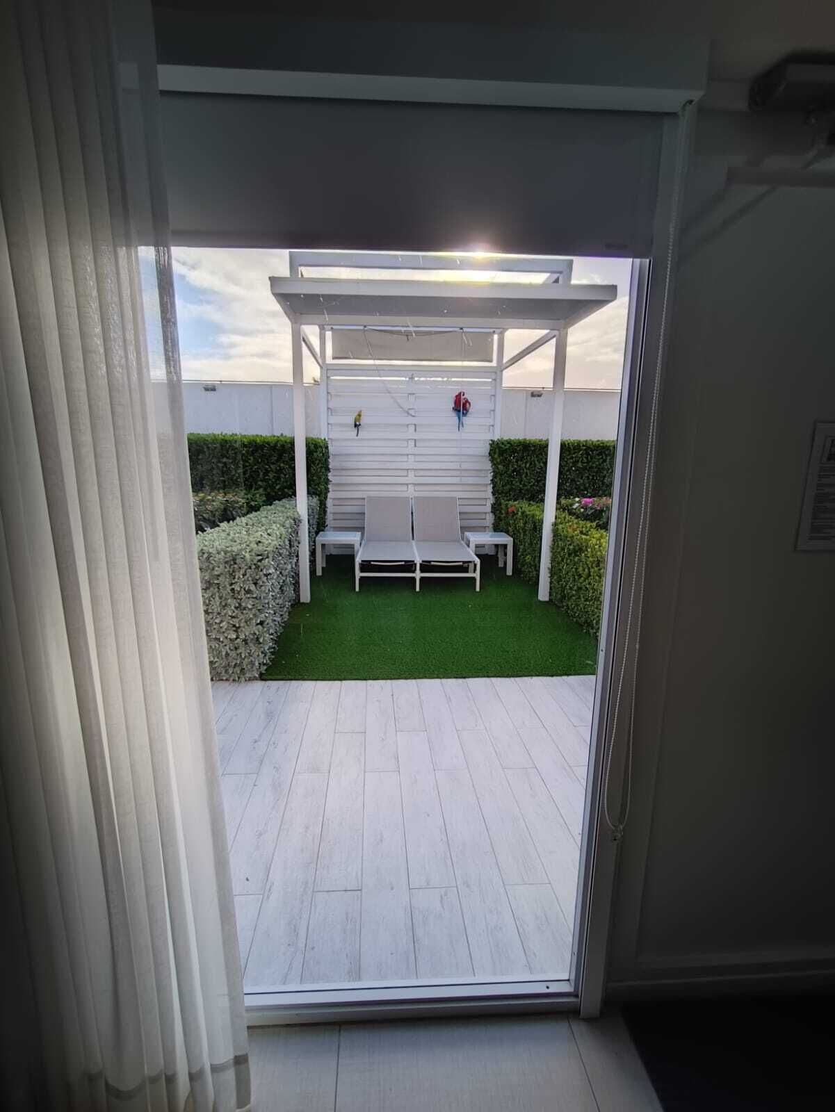 Näkymä terassialueelle, jossa on valkoinen pergola, kaksi valkoista penkkiä ja pieni pöytä.