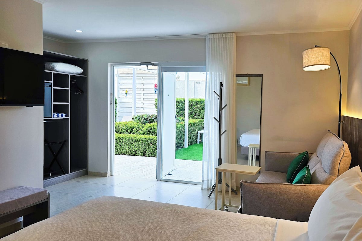 A hotel room with a bed, a desk, a chair, a wardrobe, and a balcony with a view of the garden.
