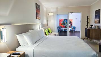 Une chambre d’hôtel avec un grand lit, une table de chevet avec une lampe, une petite table avec une chaise, et une vue sur l’extérieur par une porte vitrée.