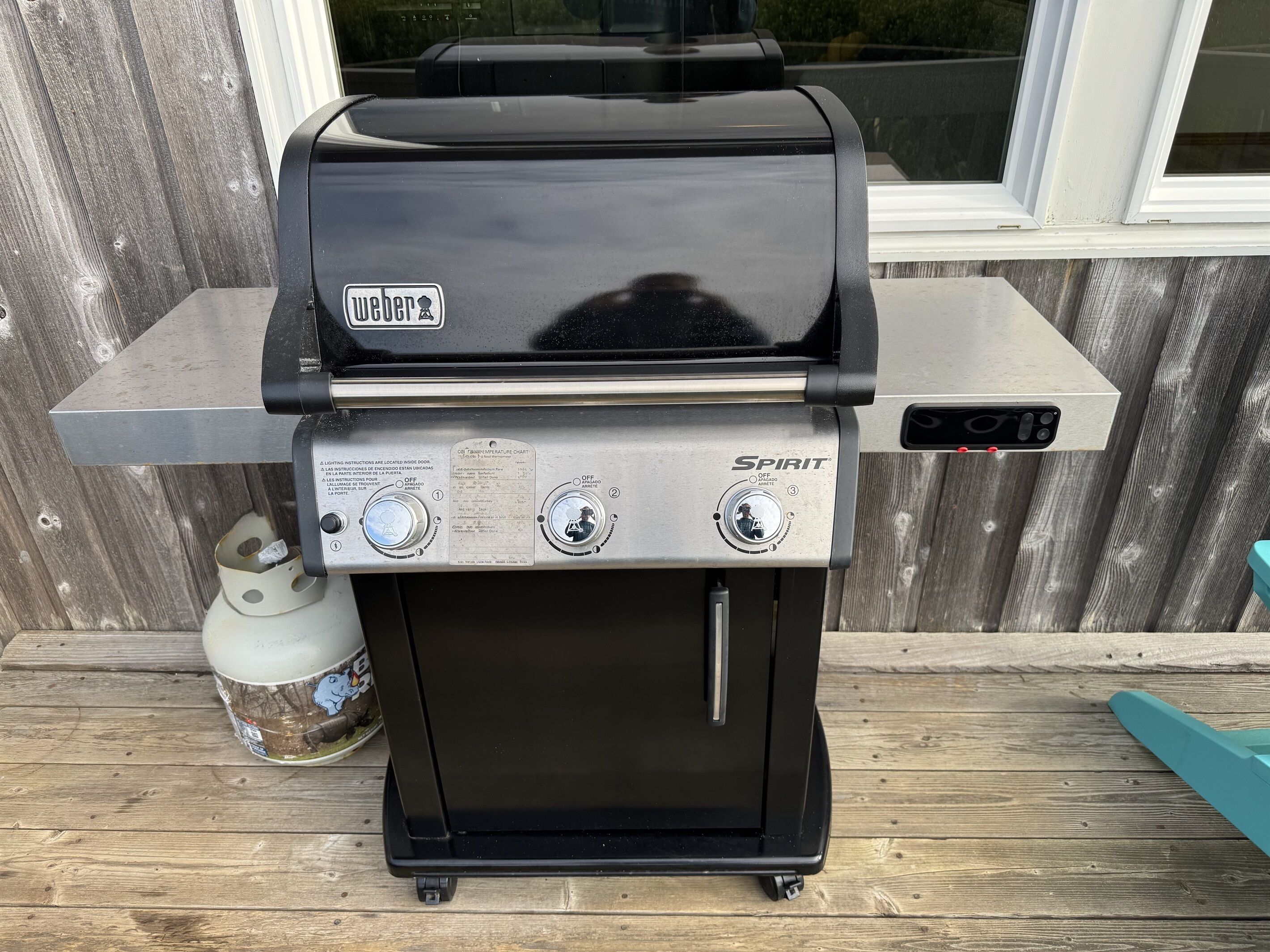Uma churrasqueira Weber Spirit preta com duas manípulos de controle e uma prateleira lateral de aço inoxidável, colocada sobre um deck de madeira.