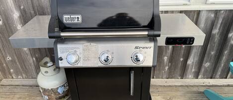 Uma churrasqueira Weber Spirit preta com duas manípulos de controle e uma prateleira lateral de aço inoxidável, colocada sobre um deck de madeira.