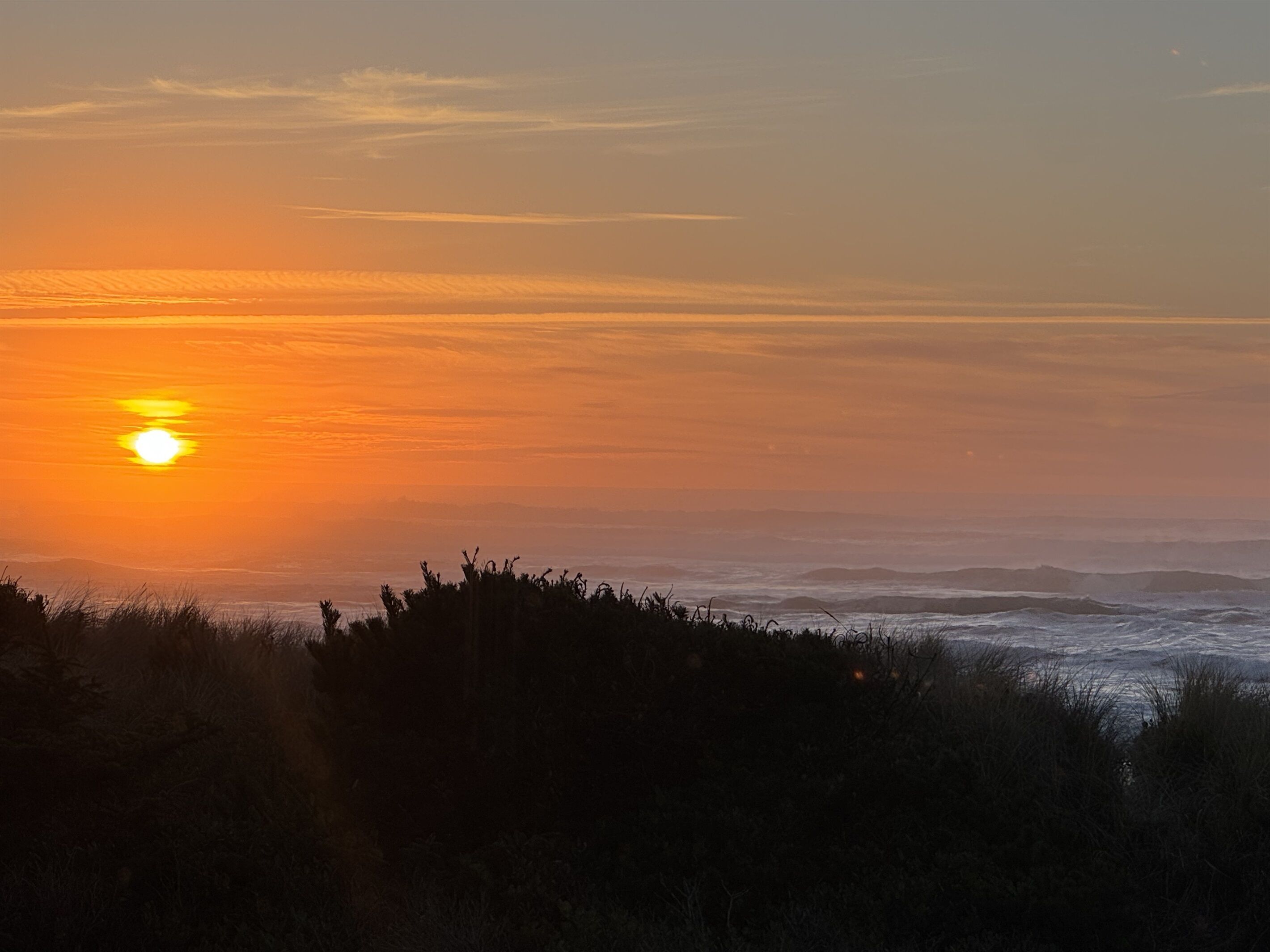 A sunset over a coastal landscape with silhouettes of vegetation and distant hills.