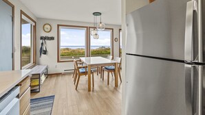 A kitchen with a large island, wooden chairs, a stainless steel refrigerator, and large windows overlooking a beach. - Luxurious & Cozy Oceanfront Escape Steps from the Sand with Sweeping Ocean Views (Rockaway Beach)