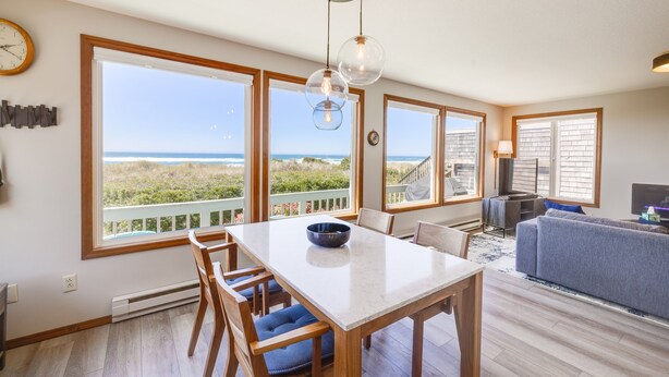 A spacious living area with a large window overlooking a beach, a dining table with chairs, and a gray sofa. - Luxurious & Cozy Oceanfront Escape Steps from the Sand with Sweeping Ocean Views (Rockaway Beach)