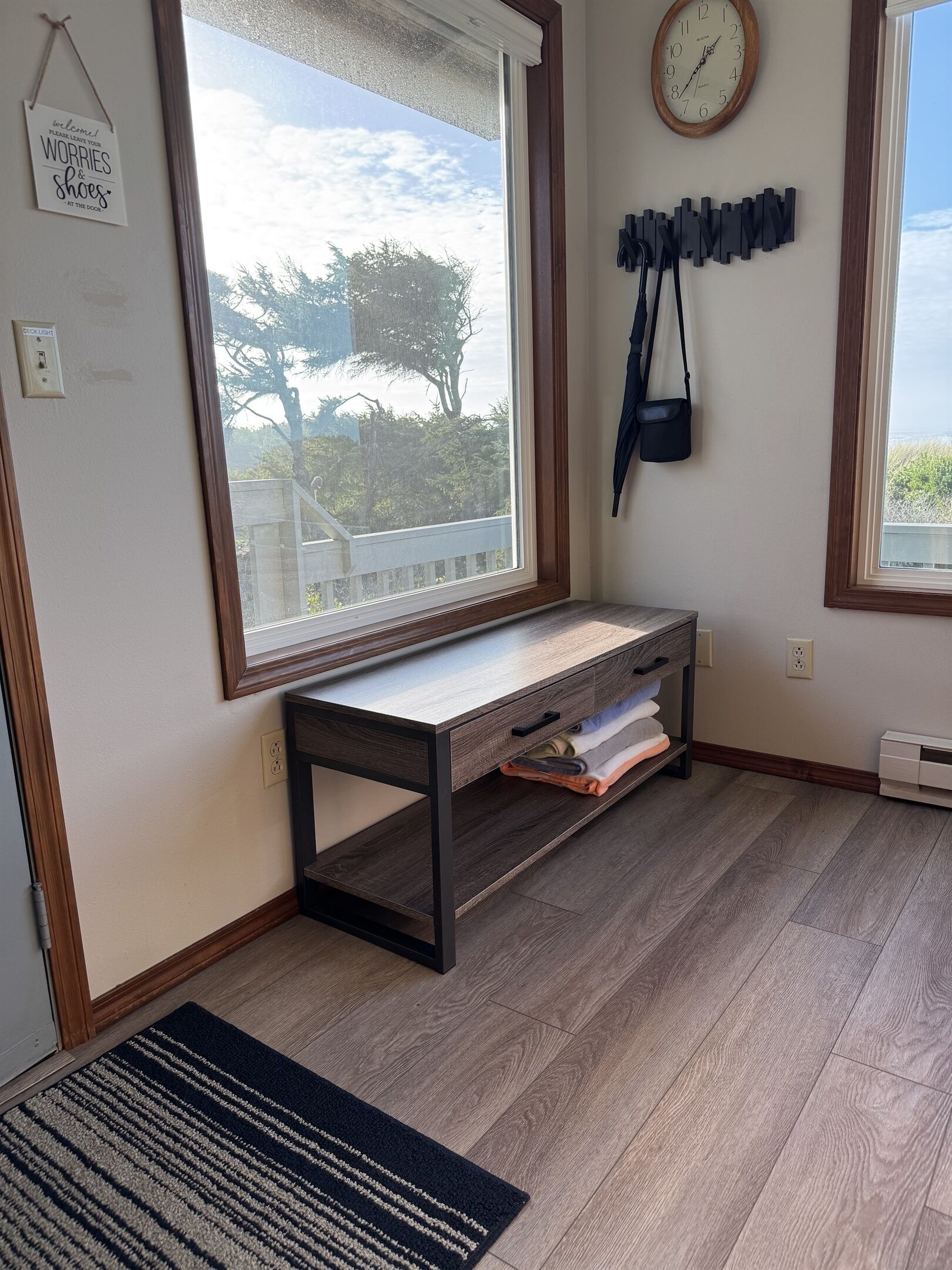A room with a large window, a wooden bench, a clock, and a wall-mounted bag rack.