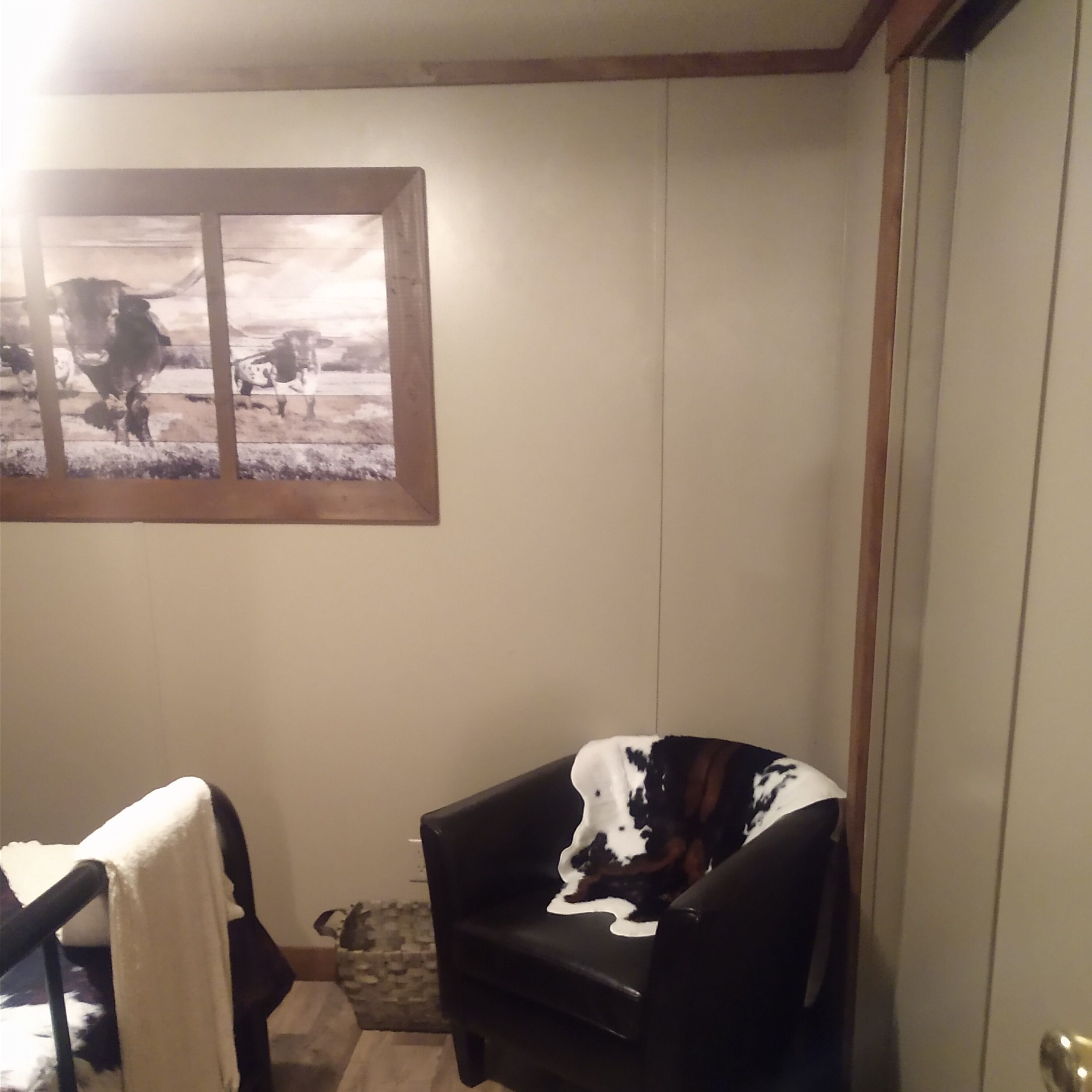 Una habitación con un cuadro enmarcado de vacas en la pared, una silla negra con una manta blanca y negra, y una pequeña alfombra en el suelo.