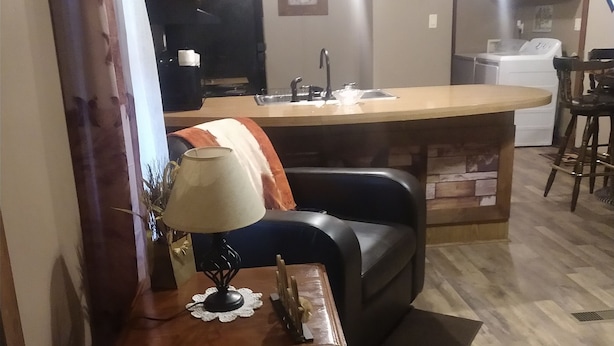 A living room with a black leather sofa, a small table with a lamp, and a kitchen area in the background. - Cozy , comfortable, and warm with unique features and plenty of amenities! (Mountain City)