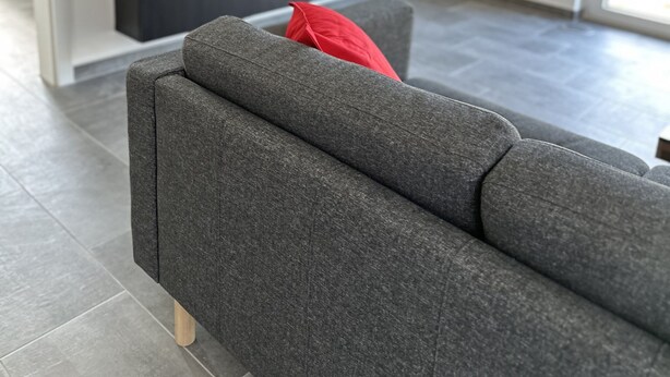 A modern living room with a grey sofa, a red pillow, and a flat-screen TV mounted on the wall displaying various channels.