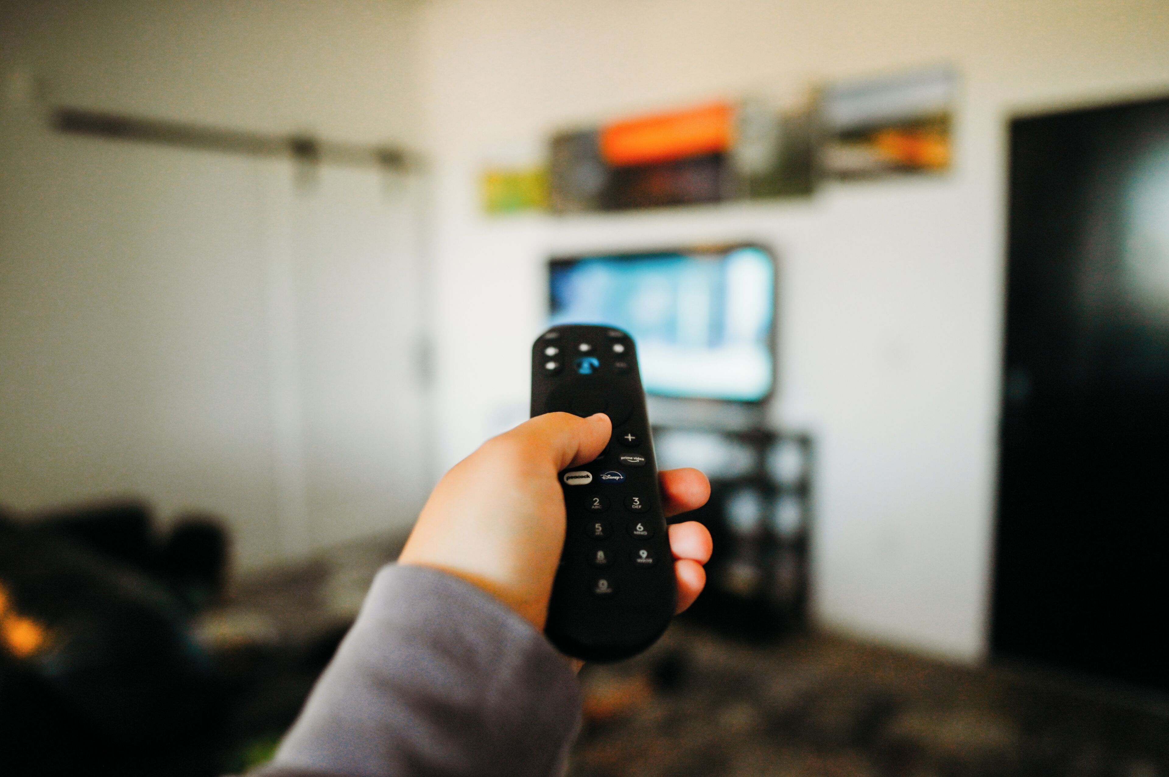 Una mano sosteniendo un control remoto en una habitación, con un televisor y una estantería al fondo.