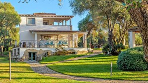 Una casa de dos pisos con balcón, rodeada de un jardín cuidado y árboles.