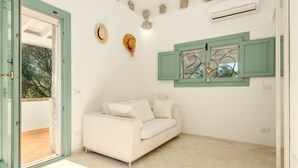 Un sofá blanco en una habitación con una puerta y contraventanas verdes, un aire acondicionado instalado en la pared, y vistas a árboles desde el exterior.