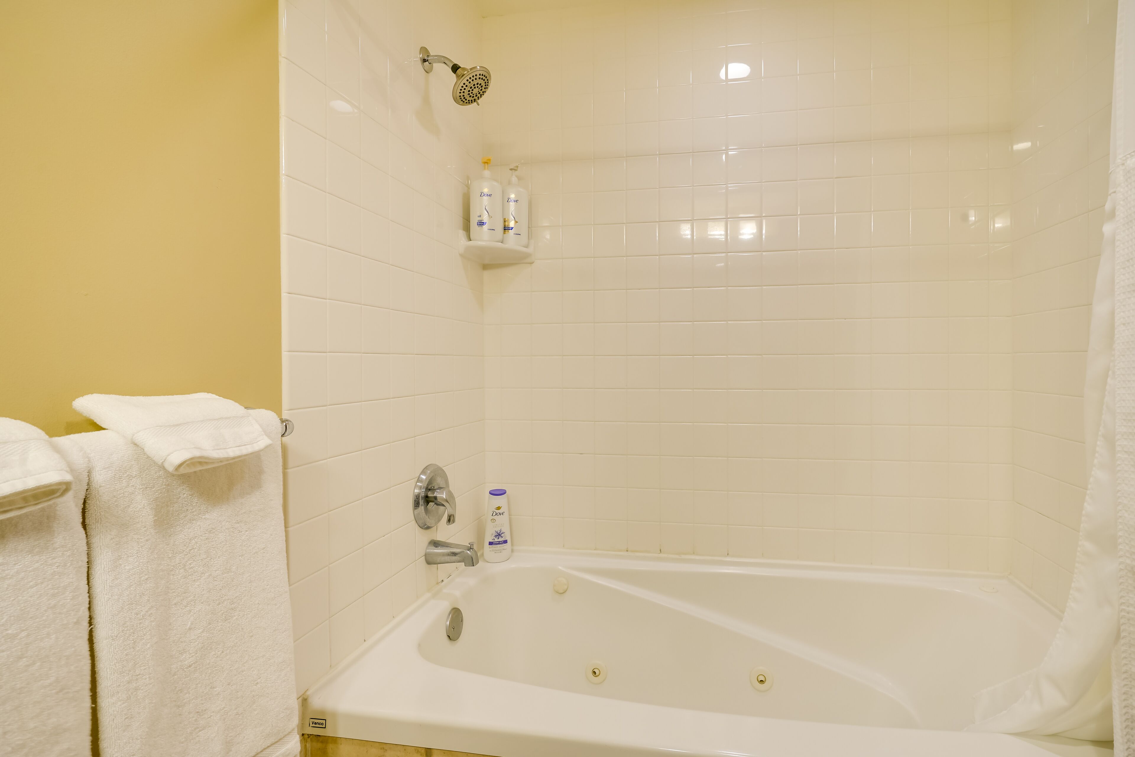 Un baño con bañera, azulejos blancos, un cabezal de ducha y dos botellas de Dove para el cuerpo en un estante.