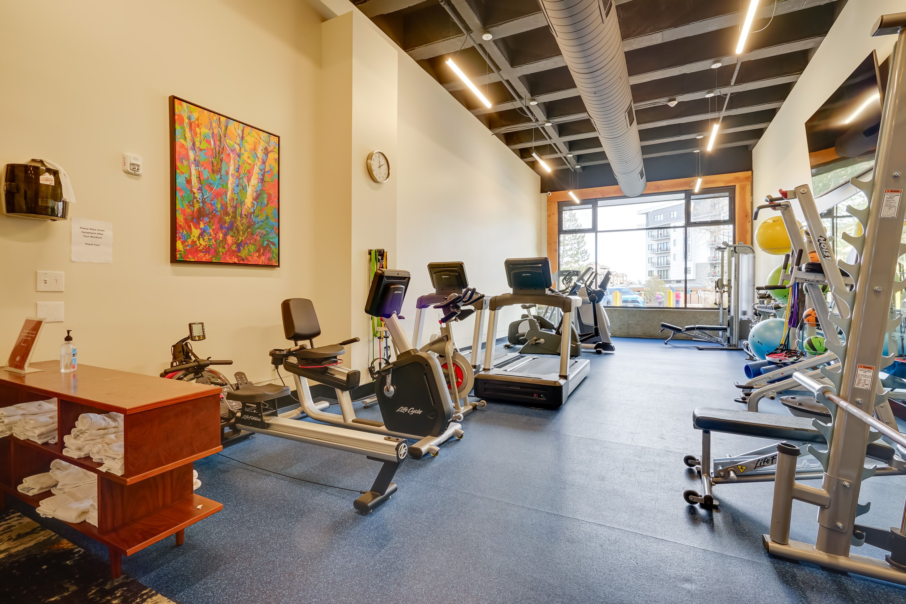 Ein Fitnessstudio mit Cardiogeräten, Krafttrainingsgeräten und einem großen Fenster mit Blick auf Gebäude.