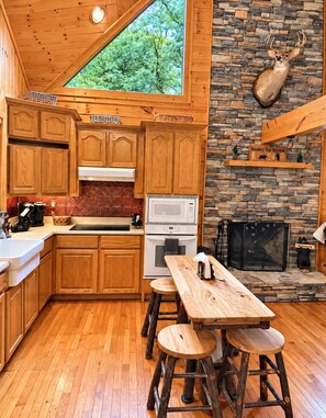 A cabin-style kitchen with wooden cabinets, a stone fireplace, and a large window overlooking greenery. - Smokey Top Retreat - 
Cozy cabin in the Great Smoky Mountains  (Robbinsville)