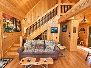 A cabin interior with a wooden staircase, a sofa with a 'Welcome to the Cabin' sign, and a rustic wooden table.